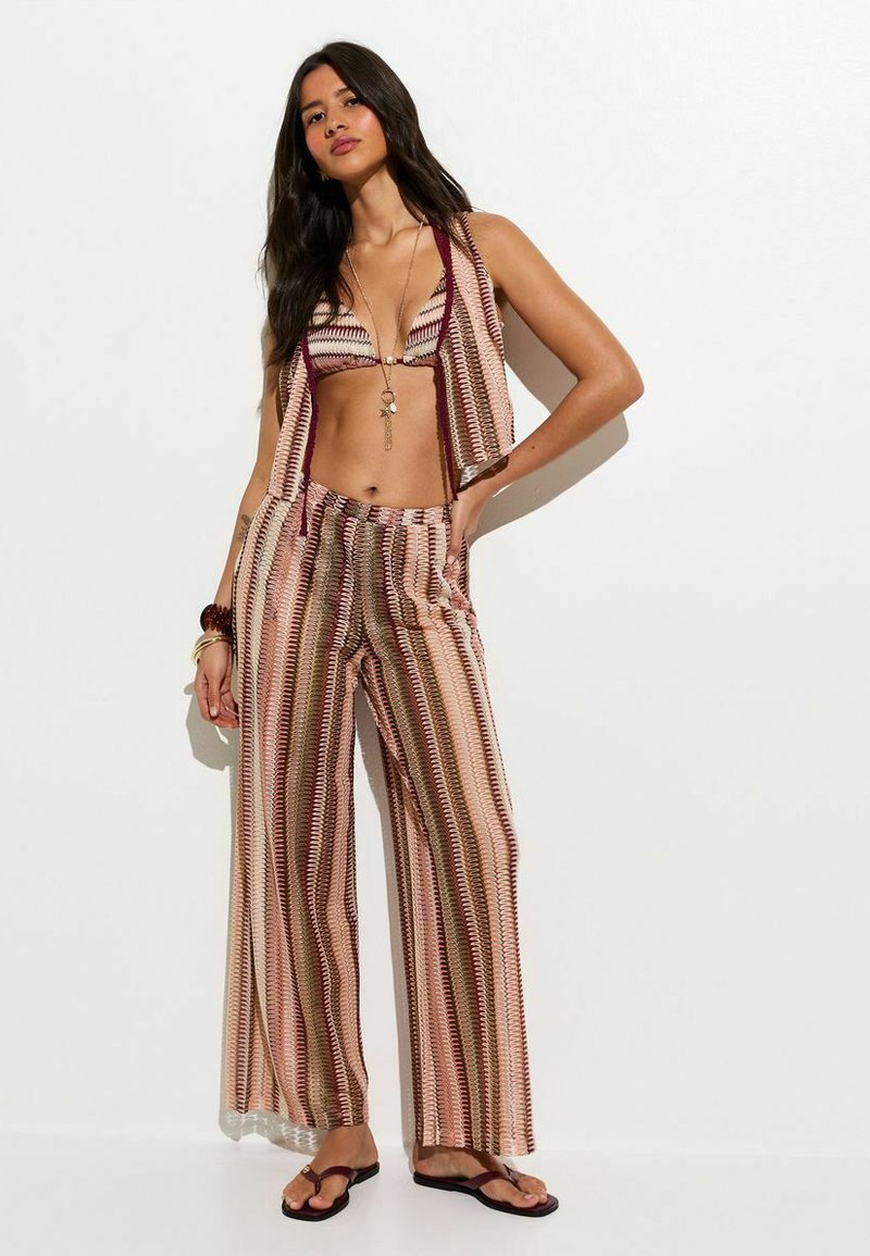 Conjunto de bikini en tonos rosa y granate con un patrón geométrico, combinado con pantalones de talle alto a juego y de corte suelto, y sandalias planas.
