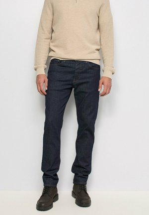 Hombre con vaqueros azul oscuro, suéter beige de manga larga y botas marrón oscuro con cordones, de pie contra una pared lisa y clara.