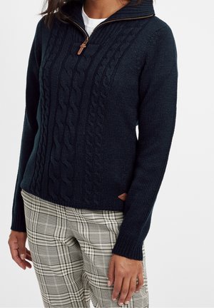 Femme portant un pull en maille torsadée bleu marine avec fermeture éclair au quart, et un pantalon beige à carreaux, debout devant un fond blanc uni.