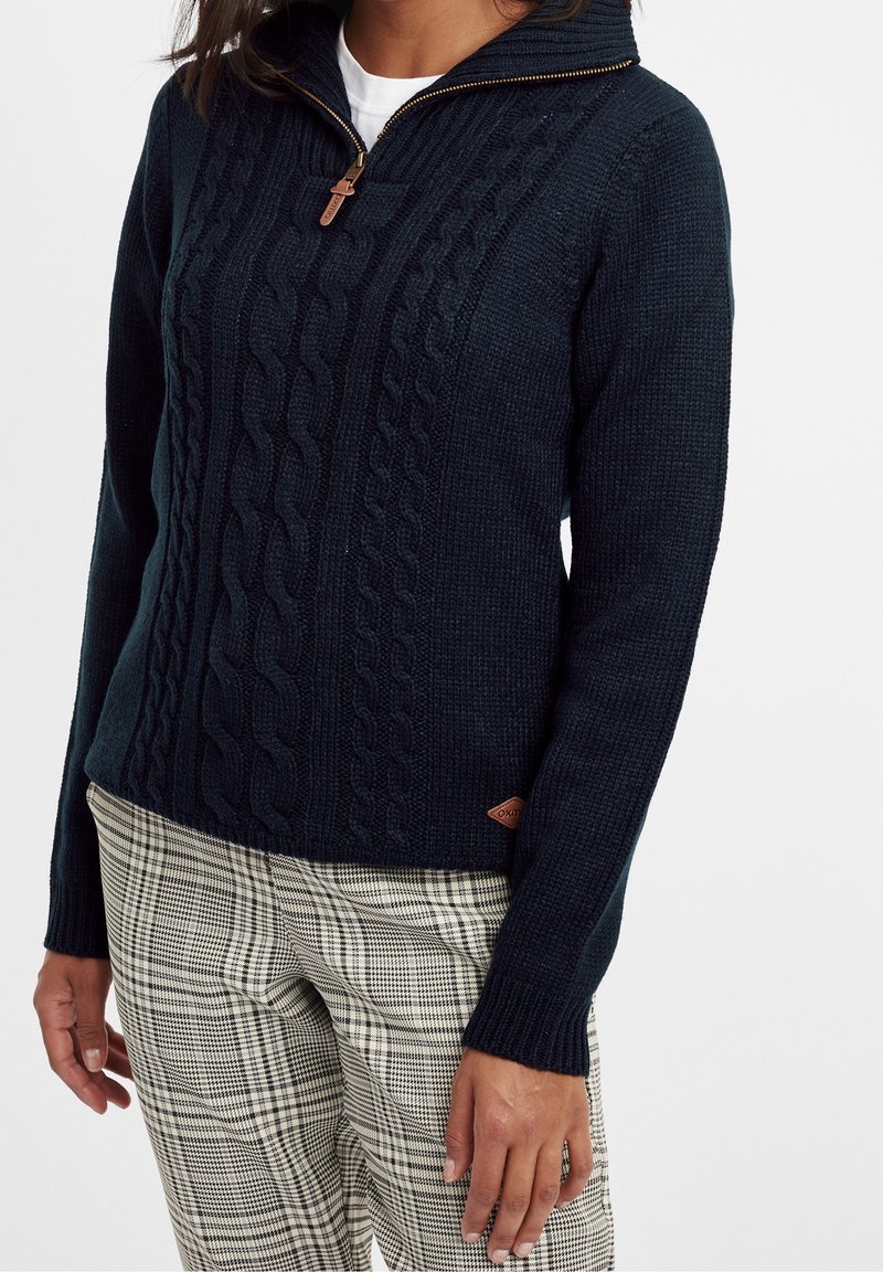 Femme portant un pull en maille torsadée bleu marine avec fermeture éclair au quart, et un pantalon beige à carreaux, debout devant un fond blanc uni.