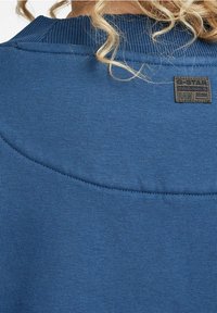 Blå sweatshirt med ribbat halsringning och sömnadsdetaljer, med en G-Star Originals etikett på övre delen av ryggen. Mjuk tygtextur.