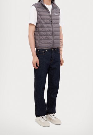 Personne portant un gilet matelassé gris, une chemise blanche à manches courtes, un jean bleu foncé et des baskets blanches, debout, les bras détendus le long du corps.