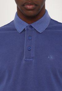 Blue polo shirt made of textured fabric featuring a pointed collar, three buttons, and a small logo on the left chest.