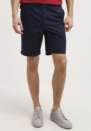 Mand iført mørkeblå shorts med diskrete prikker, en rød skjorte og lysegrå sneakers, stående mod en ensfarvet hvid baggrund.