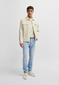 Giacca di jeans beige sopra una t-shirt grigia chiara, abbinata a jeans blu chiaro e sneakers beige. La giacca presenta due tasche sul petto.