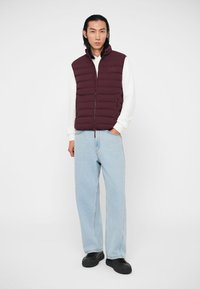 Moški v burgundskem puhovem jopiču preko belega puloverja, svetlo modrih širokih hlačah in črnih športnih copatih, stoji z eno roko v žepu.