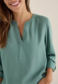 Türkise Bluse mit einem Ausschnitt, lange Ärmel, die bis zu den Ellbogen hochgerollt sind, glatte Textur und dezente Nahtdetails am Kragen.