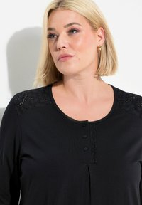 Černý top s dlouhými rukávy a krajkovými detaily na ramenou, kulatým výstřihem a zapínáním na knoflík. Měkká látka s mírným leskem a sklady v přední části.