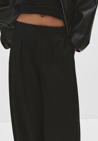 Črne hlače z krojem po meri, z nabornimi detajli, stranskimi žepi in gladko teksturo. V kombinaciji z skrajšanim črnim topom in jakno.