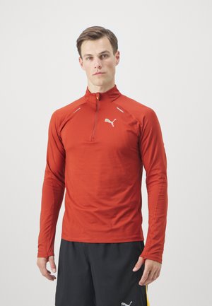 Langärmliger, mit Reißverschluss versehenen Pullover aus leuchtend rotem Stoff mit flachen Nähten und reflektierenden Akzenten, versehen mit einem kleinen weißen Logo auf der Brust.