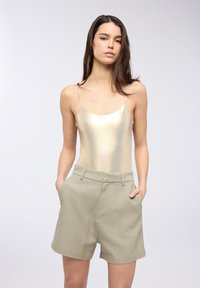Canotta metallica oro con spalline sottili e pantaloni corti verdi chiari su misura con tasche laterali e linee pulite, posizionata su uno sfondo neutro.