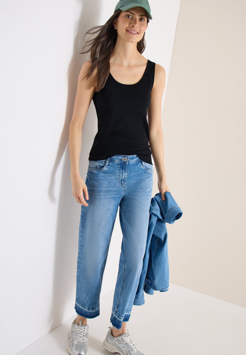 Femme portant un débardeur noir, un jean court bleu clair, une casquette verte, des baskets blanches, tenant une veste en jean bleue devant un mur bicolore.