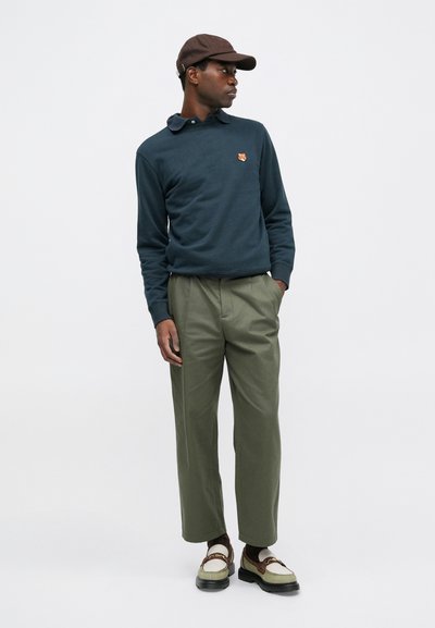 Tricou polo cu mâneci lungi de culoare bleumarin, cu guler și un emblemă mică, pantaloni largi de culoare verde măslin, o șapcă maro și pantofi bicolori.