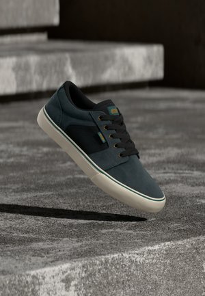 Baskets bleu sarcelle foncé et noir avec une semelle blanche flottant au-dessus d'escaliers en béton dans un cadre urbain extérieur.
