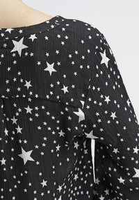 Blouse noire en tissu texturé, ornée d'un motif d'étoiles blanches, avec un col arrondi et un léger effet froncé au dos.