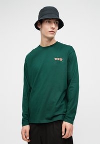 Młody mężczyzna w ciemnozielonej koszuli z długim rękawem z małym czerwonym napisem i wzorem pałeczek, w czarnych spodniach i czarnym kapeluszu typu bucket.