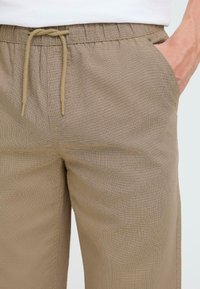 Beige drawstring pants with an elastic waist, featuring a textured fabric pattern and side pockets. Smooth finish with a relaxed fit.