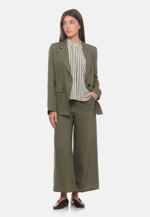 Giovane donna che indossa un completo verde oliva con pantaloni a gamba larga e una camicia a righe con bottoni, in piedi con una mano in tasca e guarda di lato.