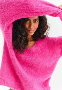 Miękki, oversize różowy sweter z szerokim dekoltem i długimi rękawami, o delikatnej fakturze, z tkanymi metkami na dole.