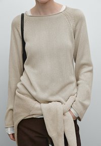 Beige gebreide trui met een brede halslijn en lange mouwen, gelaagd over een wit shirt. Losse pasvorm met een knoopdetail rond de taille.