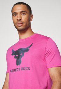 Mann iført en lys rosa T-skjorte med svart okse-logo og teksten "Project Rock", som ser litt til siden mot en ensfarget bakgrunn.