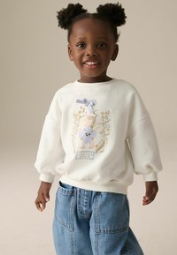 Weißes Sweatshirt mit gepufften Ärmeln, das ein farbenfrohes Blumenmotiv und einen lavendelfarbenen Schleifenakzent zeigt. Kombiniert mit blauen, weiten Jeans aus Denim.
