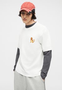 Νεαρό άτομο που φοράει λευκό μπλουζάκι με μικρό γραφικό Charmander, γκρι μακριά μανίκια, γκρι παντελόνι και κόκκινο καπέλο με φθορές.