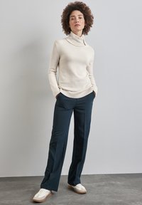 Maglione a collo alto di colore crema con orlo a coste, abbinato a pantaloni a gamba larga blu navy. Scarpe in evidenza sono sneakers bianche con dettagli beige.