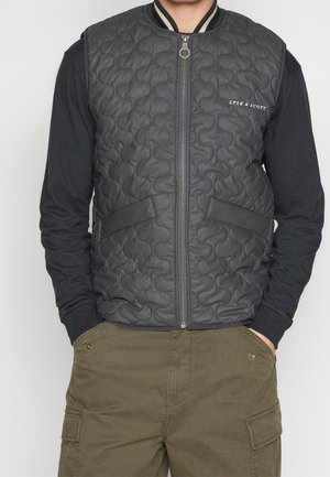 Homme portant un gilet matelassé noir zippé avec le logo Lyle & Scott sur un t-shirt noir à manches longues et un pantalon cargo vert olive.