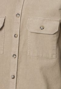 Close-up van een beige corduroy shirt met flapzakken en gemarmerde knopen aan de voorkant.