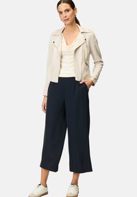 Beige Kunstlederjacke mit Reißverschlüssen, über einem hellen Creme-Oberteil mit Schalkragen und marineblauen weiten Hosen getragen, kombiniert mit weißen Sneakers.