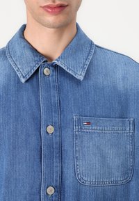 Lys blå denim skjorte med knapper. Har spiss krage, sølvfargede knapper, og en venstre brystlomme med rød, hvit og blå logo broderi.
