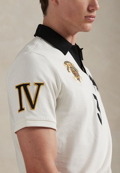 Λευκό πόλο πουκάμισο με μαύρο γιακά, που διαθέτει κεντημένες λεπτομέρειες με χρυσές αποχρώσεις και μεγάλο μαύρο αριθμό "3" στο μπροστινό μέρος και "IV" στο μανίκι.