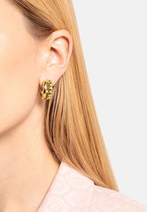 Goldene Creolen haben eine glatte, polierte Oberfläche mit ineinander verwobenen Designs und runden Akzenten. Die Ohrringe sind neben einem hellrosa gemusterten Stoff präsentiert.