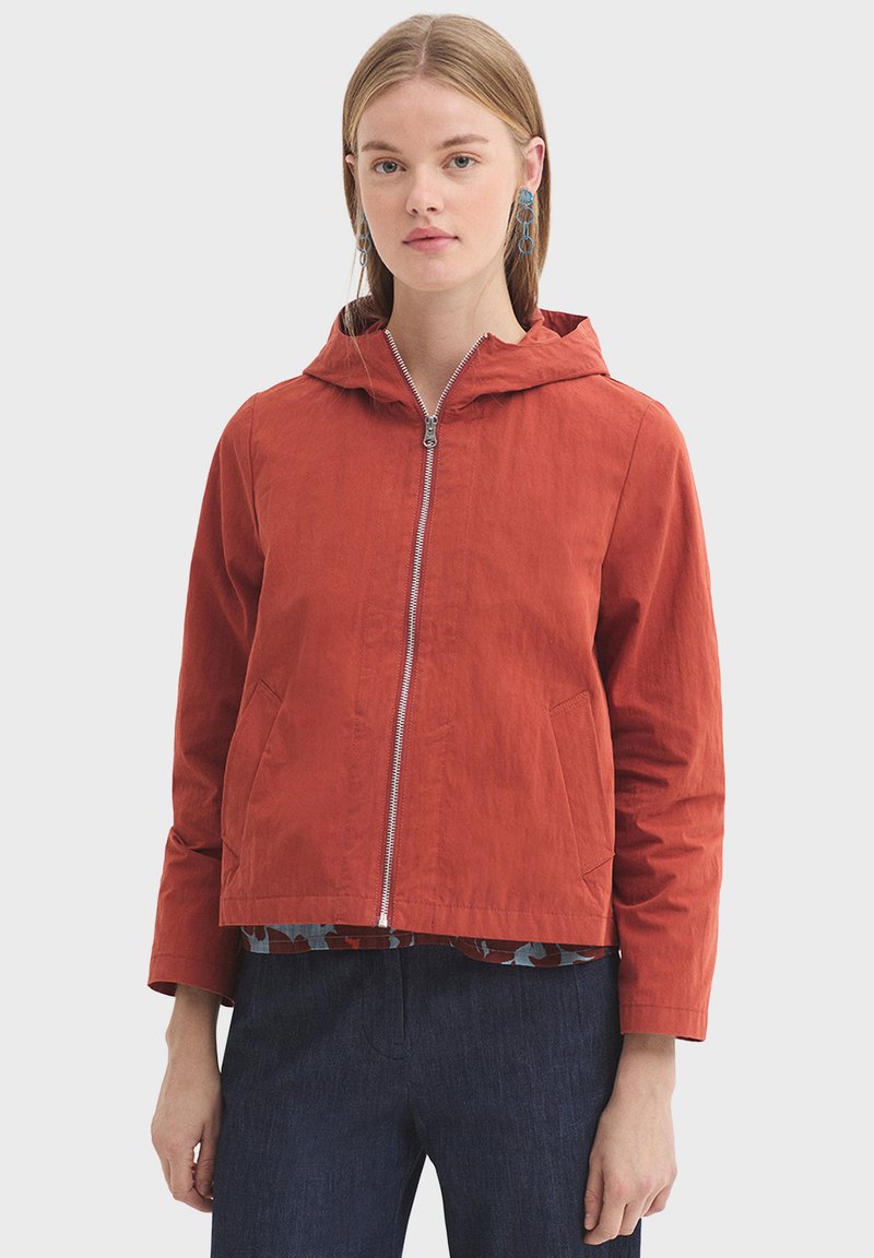 Veste à capuche rouge avec fermeture éclair frontale, poches latérales, et texture légère. Présente une doublure intérieure multicolore.