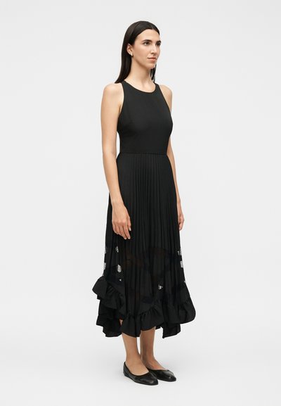 Γυναίκα που στέκεται φορώντας ένα αμάνικο μαύρο φόρεμα με πιετέ φούστα και βολάν στο τελείωμα, σε συνδυασμό με μαύρα φλατ παπούτσια, σε απλό λευκό φόντο.