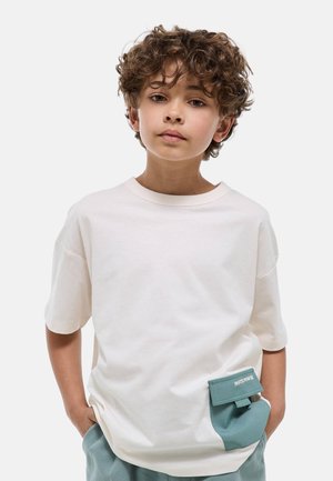 Garçon aux cheveux bouclés portant un T-shirt blanc avec une poche turquoise et un pantalon turquoise, debout les mains dans les poches devant un fond uni.