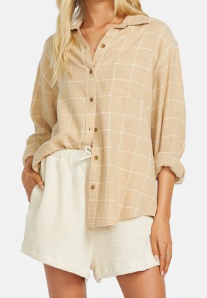 Nainen pukeutuneena beige ruudulliseen nappipaitaan käärittyine hihoineen ja valkoisiin tekstuurillisiin shortsseihin, käsi taskussa, kasvot eivät näy.