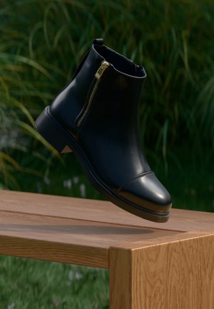 Bottine noire en cuir avec fermeture éclair dorée flottant au-dessus d'un banc en bois à l'extérieur avec un fond de feuillage vert.