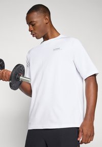 Camiseta deportiva blanca de manga corta con cuello redondo y un pequeño logo gris. El modelo sostiene una mancuerna negra y lleva pantalones cortos oscuros.