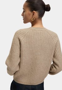 Mujer con un peinado de moño trenzado que lleva pendientes gruesos de aro de oro y un suéter de punto texturizado beige, de perfil contra un fondo blanco.