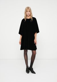 Rochie din catifea neagră cu mâneci scurte și largi și o croială lejeră. Modelul poartă ciorapi negri și pantofi negri cu vârf ascuțit, stând în fața unui fundal neutru.