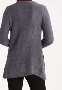 Osoba v šedém pleteném kardiganu s dlouhým rukávem a mírně asymetrickým lemem, v černých kalhotách, sledována zezadu.