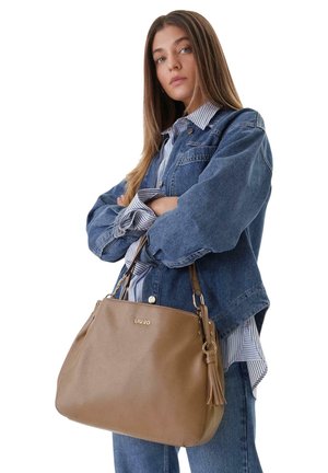 Joven mujer con chaqueta vaquera y camisa de rayas sostiene un gran bolso marrón Liu Jo con detalle de borla, brazos cruzados.