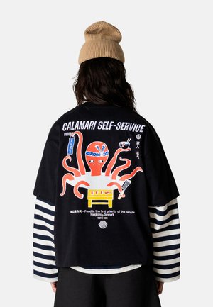 Person iført en sort skjorte med en farverig blækspruttegrafik, som holder mad og en kødøkse, teksten på ryggen lyder "Calamari Self-Service".