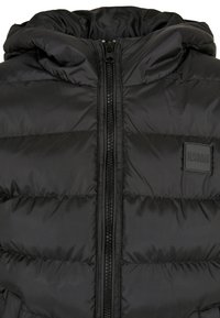 Veste matelassée noire avec fermeture éclair à l'avant, capuche attachée, coutures horizontales et une petite étiquette "Urban Classics" sur la poitrine.