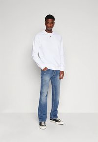 Valge pusa ümmarguse kaelusega, ühe punase logoga, kombineeritud heleroheliste teksadega; modell kannab must-valgeid tossusid.