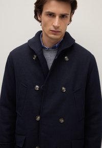 Abrigo cruzado de mezcla de lana azul marino con botones plateados, cuello alto y bolsillos laterales. Suéter gris debajo con camisa de rayas azules.