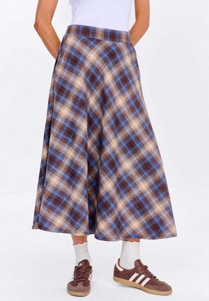 Kobieta ubrana w brązową i niebieską kraciastą spódnicę midi, białą bluzkę, białe skarpetki i brązowe sneakersy Adidas, stojąca na jednolitym tle.