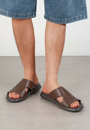 Jambes portant un short en jean bleu et des sandales à enfiler ouvertes aux orteils de couleur marron foncé sur un sol gris contre un mur blanc.